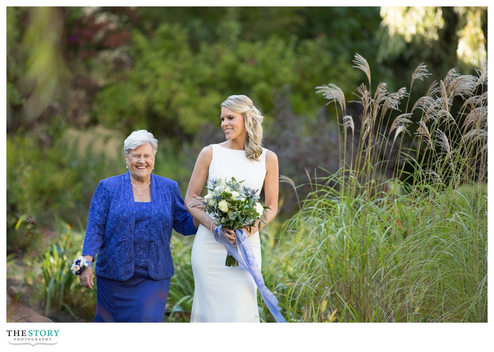 bride and her mother walk path toward wedding ceremony on bridge at Mirbeau